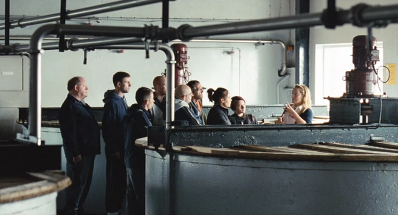 The Angel’s Share (2012) movie still: wide — A spacious industrial facility features large vats and exposed metal piping overhead. A blonde woman…