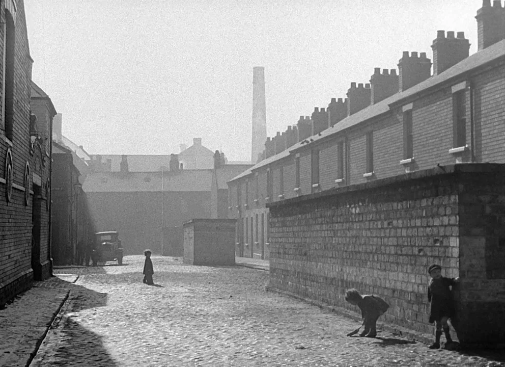 Odd Man Out (1947) movie still: wide — A row of industrial brick terraced houses lines a narrow, hazy cobblestone street. Three small children…