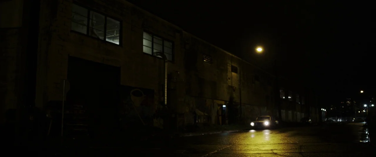 American Mary (2012) movie still: wide — A dark, desolate industrial area features a large concrete building with illuminated windows at night. A…