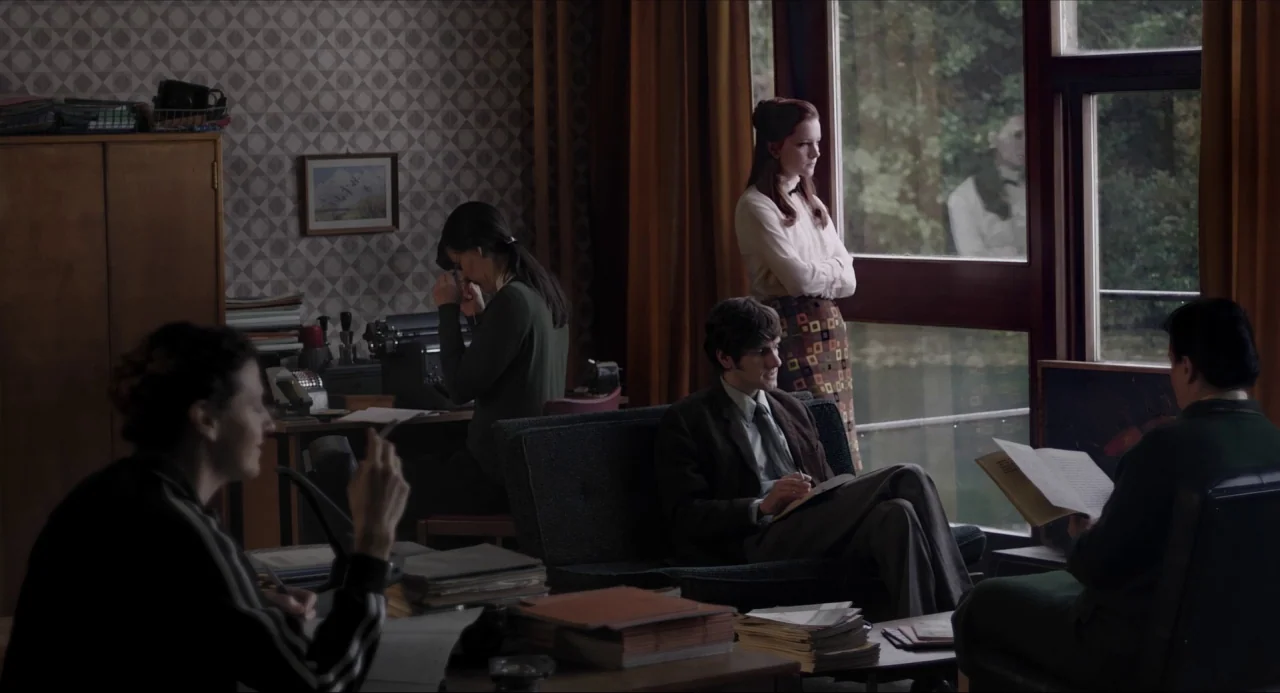The Falling movie still: wide — A 1970s office with wallpapered walls and multiple desks cluttered with stacks of papers. Four individuals are…