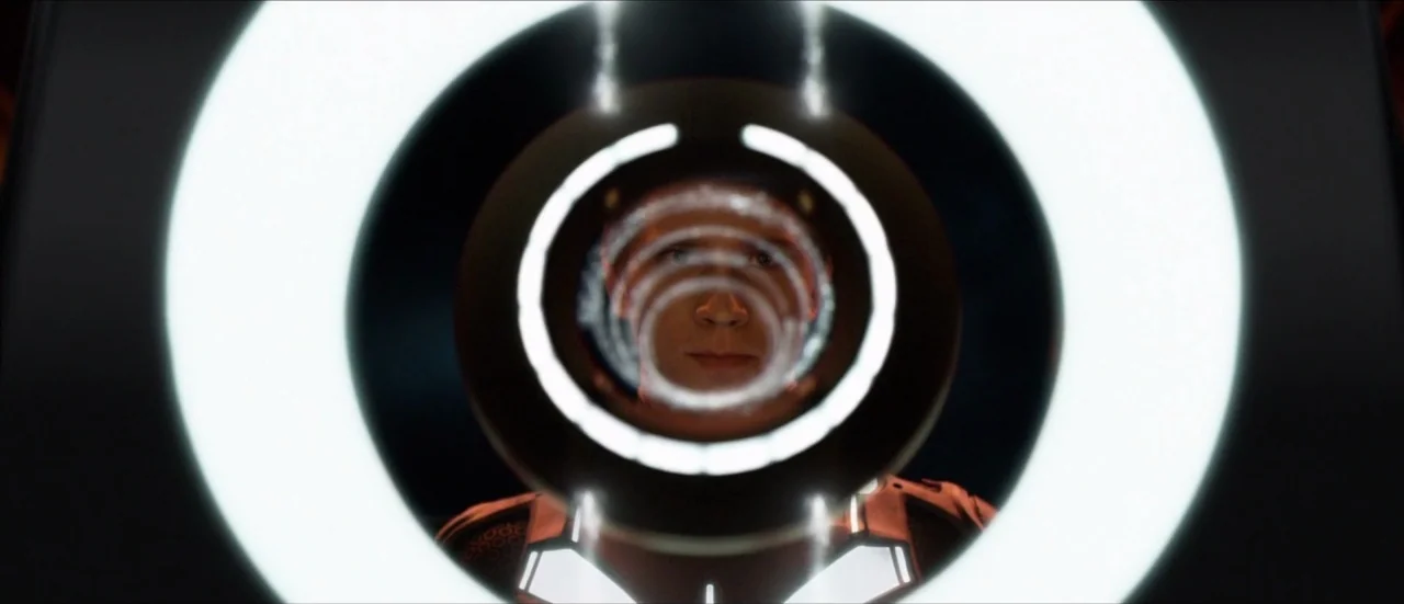 TRON: Legacy (2010) movie still: closeup — A dark interior is dominated by a large, glowing white ring-shaped aperture. A young man gazes through…
