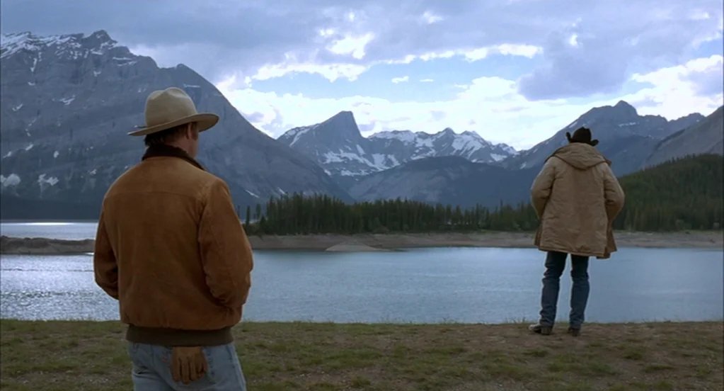 Brokeback Mountain (2005) movie still: fullbody — A vast alpine lake sits before a range of towering, snow-capped rocky mountains. Two men stand…