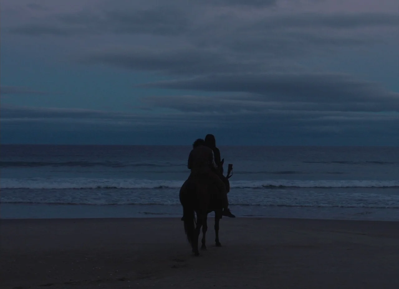 The Nightingale (2018) movie still: wide — A flat stretch of dark beach meets the churning ocean under a cloudy dusk sky. Two people ride a horse…