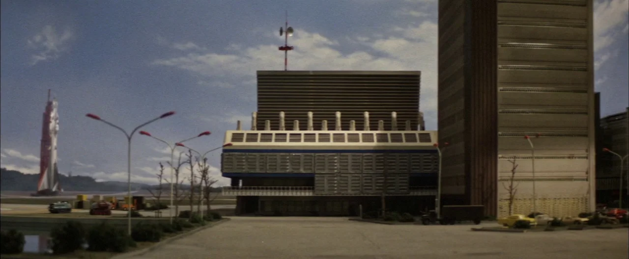 Destroy All Monsters (1968) movie still: establishing — A miniature-style model of a concrete industrial facility stands beside a tall tower. Sparse…