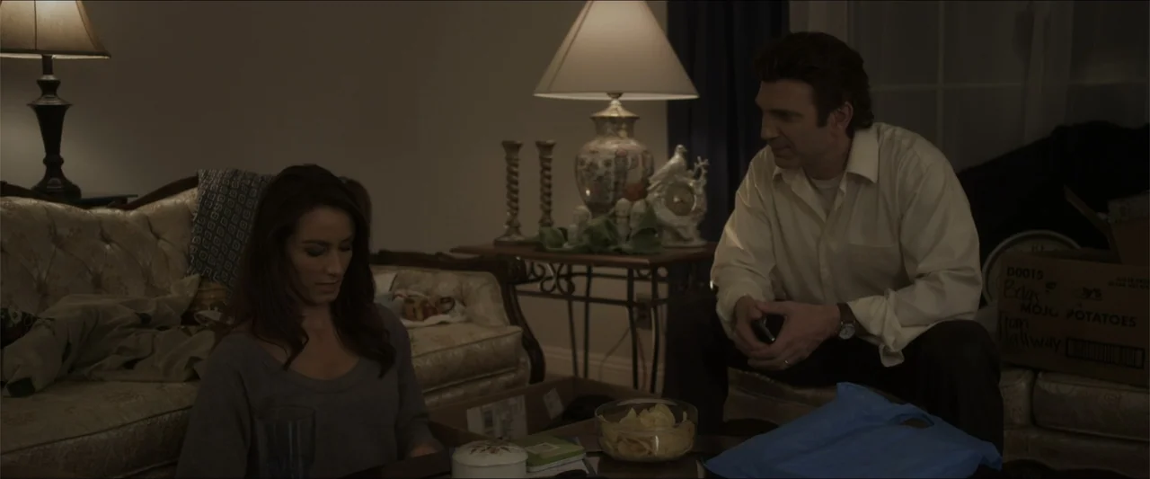 Heavenly Deposit (2019) movie still: medium — A living room interior contains a sofa, moving boxes, and table lamps at night. A woman in a grey…