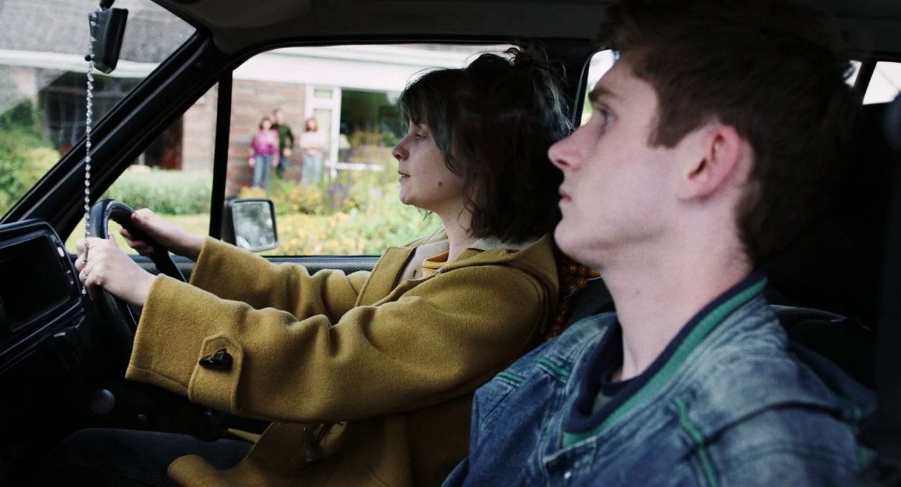Dating Amber movie still: medium — Inside a car, a young woman steers the wheel while a man sits beside her. Outside the driver's window, a house…