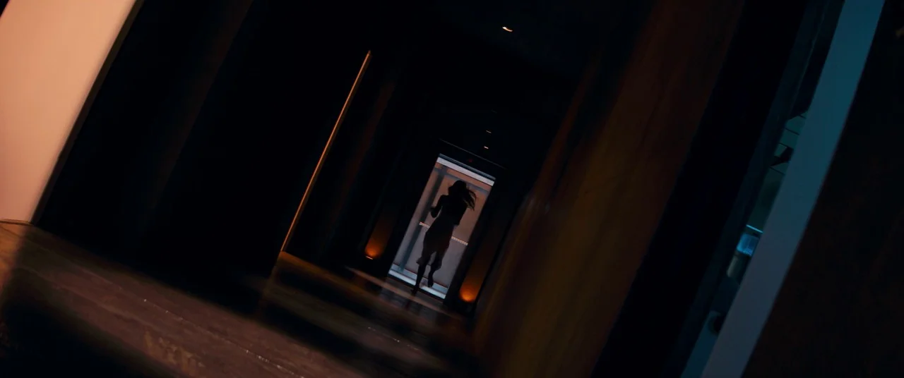 dutch angle shots example from Elizabeth Harvest: A dark, narrow corridor with warm-lit recesses leads to a brightly lit doorway. A woman is silhouett