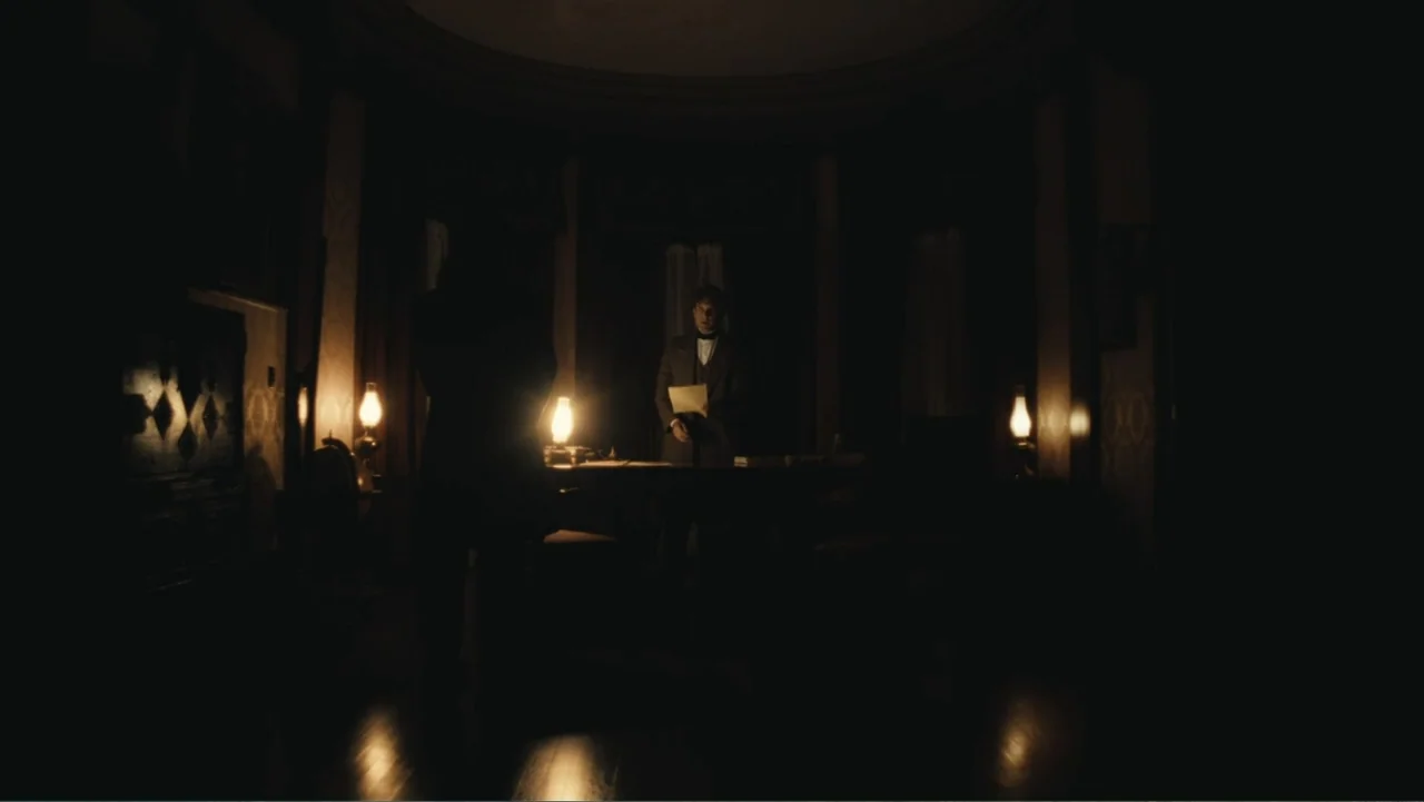 Demon in the White House (2021) movie still: medium — A dimly lit 19th century room contains a dark wood desk and oil lamps. One man stands by the…