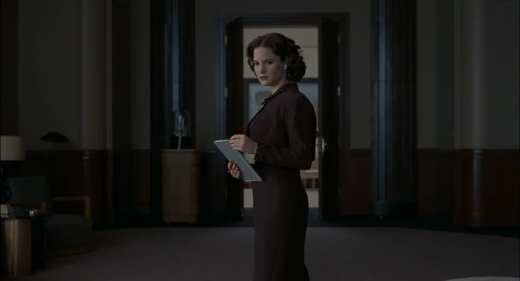 The Hudsucker Proxy (1994) movie still: medium — A dark, wood-paneled space features high ceilings and a distant open double doorway. A woman stands…
