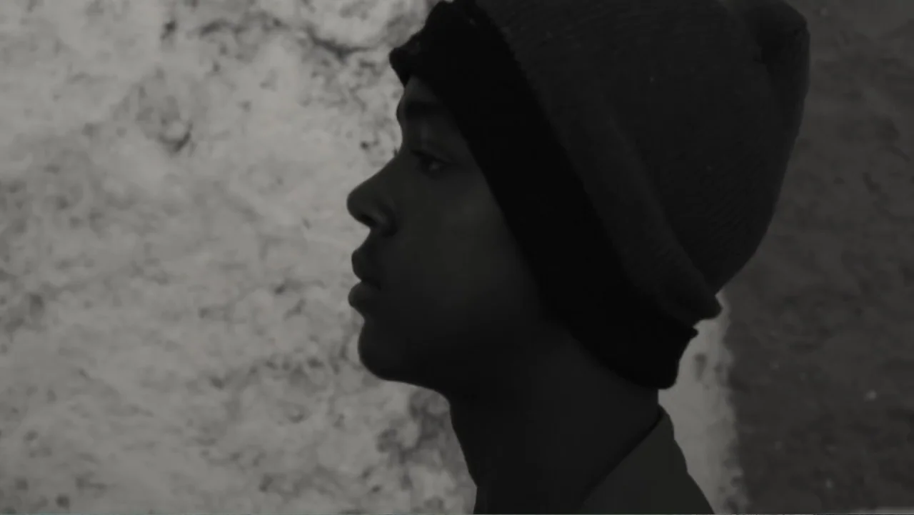 Faya Dayi (2021) movie still: closeup — An outdoor urban location features a textured, light-colored concrete wall behind the subject. A young man…