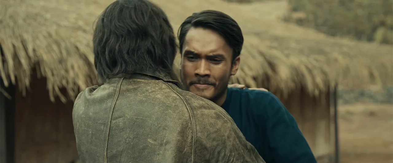 The Unholy (2022) movie still: medium — A rural setting features a hut with a thatched roof in the background. One man with dark hair wears a…