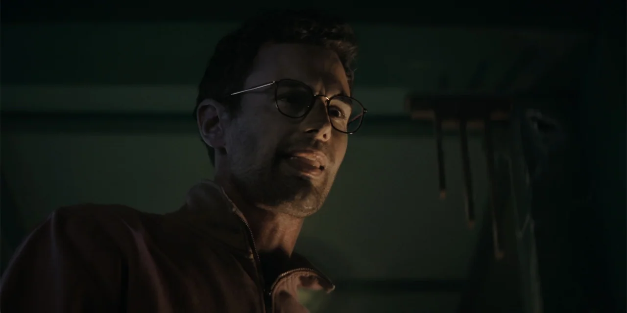 The Monkey (2025) movie still: closeup — A dimly lit room provides the backdrop for a man speaking into the shadows. The man gazes downward while…