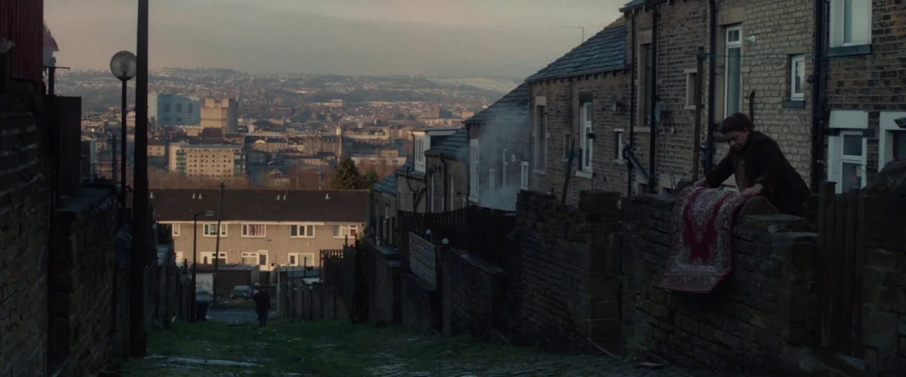 Ali & Ava (2021) movie still: wide — A steeply sloped residential alleyway features brick homes and a view of the city. A woman stands by a stone…