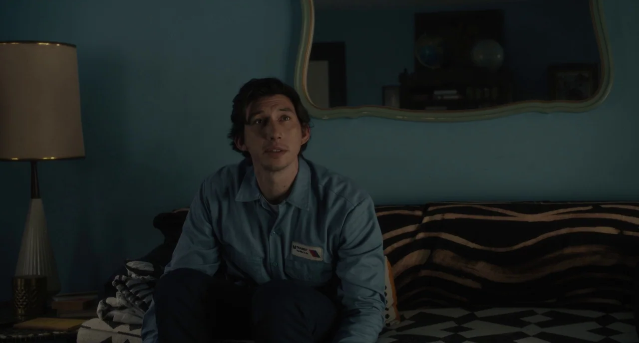 Paterson (2016) movie still: medium — A dimly lit room features a teal wall with an ornate hanging mirror above. A man in a blue uniform sits on a…