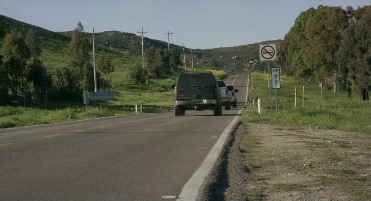 Instantes Capturados (2022) movie still: wide — An asphalt road winds through green hills under a clear bright sky. Three dark vehicles travel along…