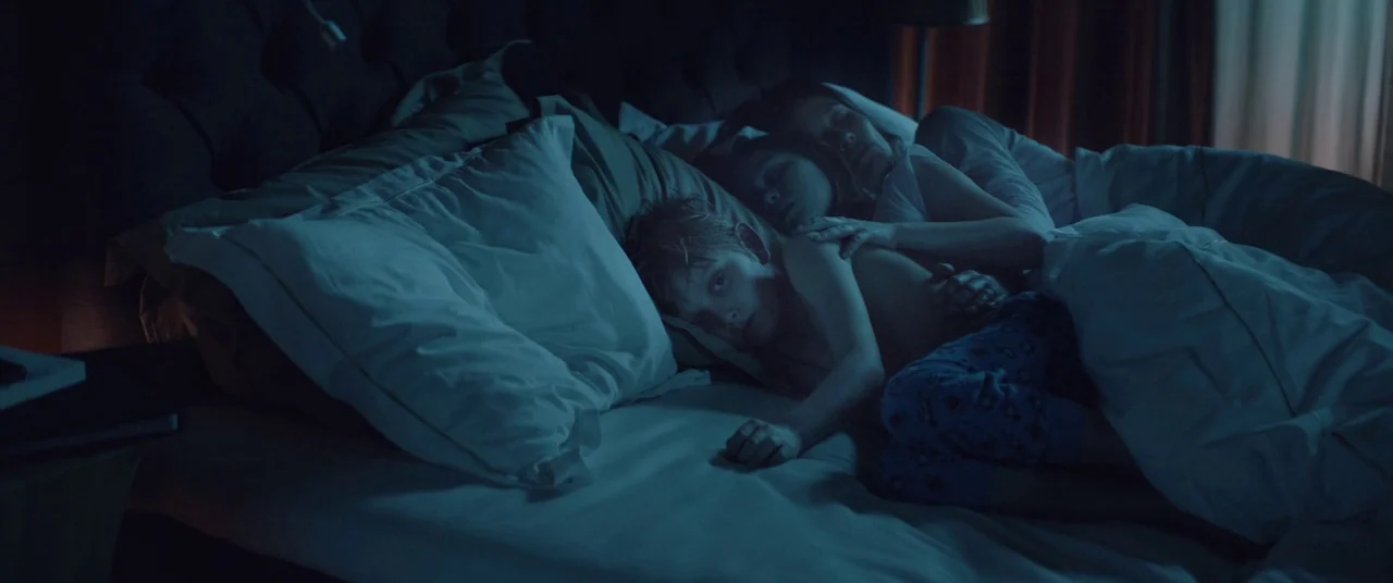 Force Majeure (2014) movie still: medium — A dark bedroom shows a child and woman lying together on a large bed. The child gazes toward the camera…