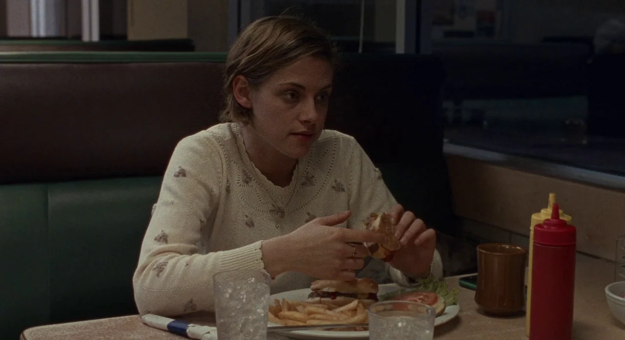 Certain Women (2016) movie still: medium — A quiet diner booth at night with dark green vinyl upholstery and laminate tables. A young woman in a…