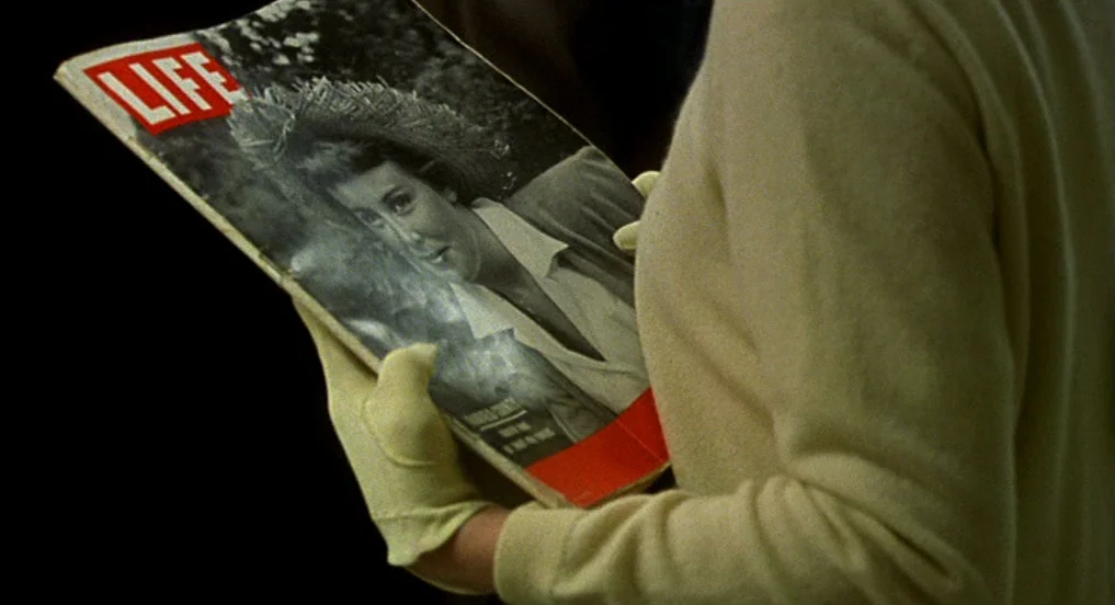 Pollock (2000) movie still: closeup — A close view shows a person in a beige sweater holding a Life magazine. The subject wears yellow gloves while…