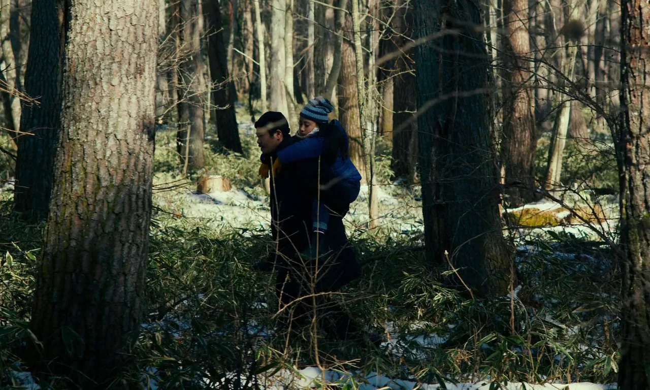 Evil Does Not Exist (2023) movie still: fullbody — A dense woodland area with tall trees, patches of snow, and low-growing ground cover. A man walks…