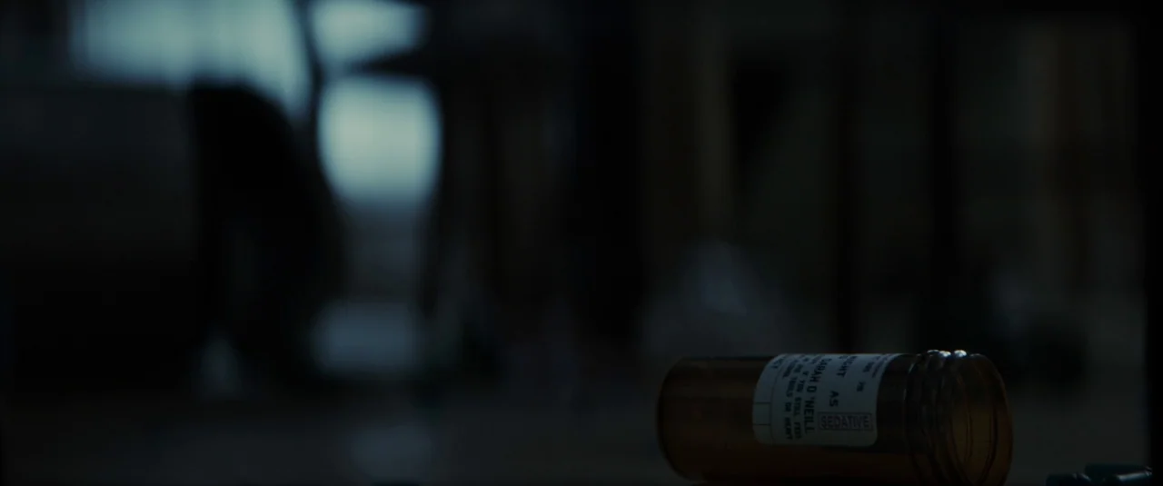 The Hole in the Ground (2019) movie still: closeup — An orange prescription bottle lies horizontally on a dark surface in a shadowed room. The label…