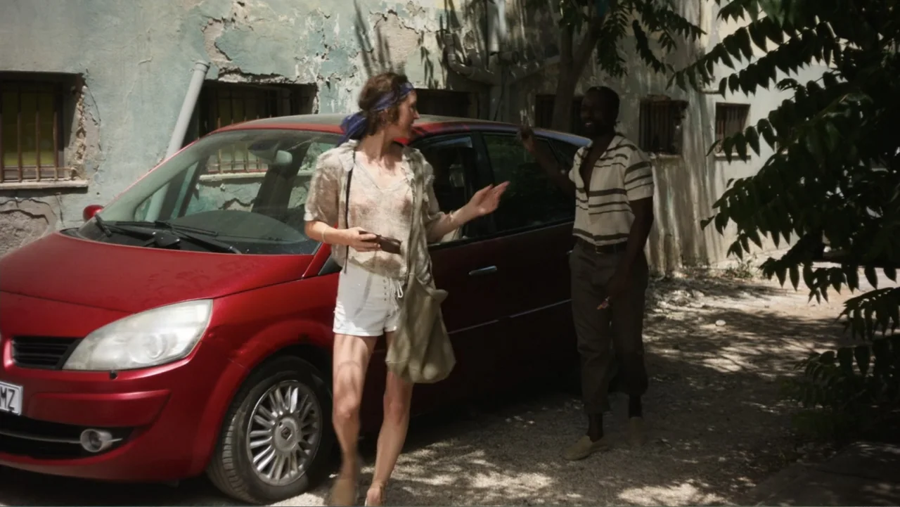 Hot Milk (2025) movie still: fullbody — An outdoor area beside a weathered building wall with a red hatchback parked nearby. A woman and man stand…