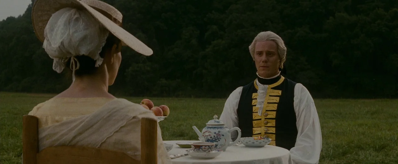 The Last of the Mohicans (1992) movie still: medium — Two individuals sit at a small table covered in white linen in a grassy field. A painted…