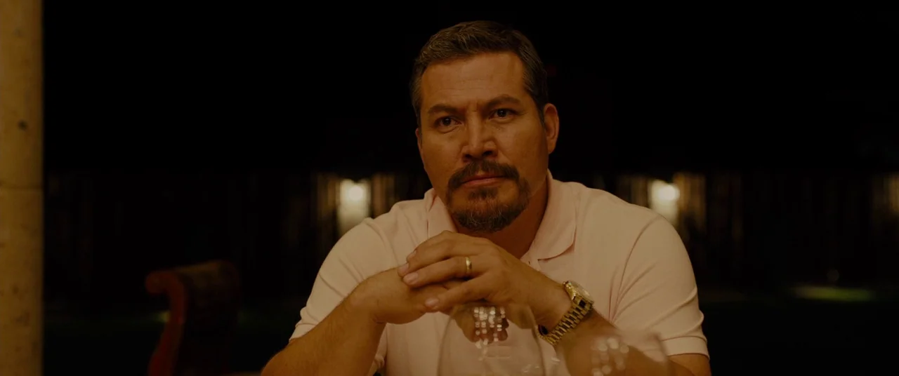 Sicario (2015) movie still: medium — A dark outdoor space with indistinct background lighting at night. A middle-aged man sits at a table with his…