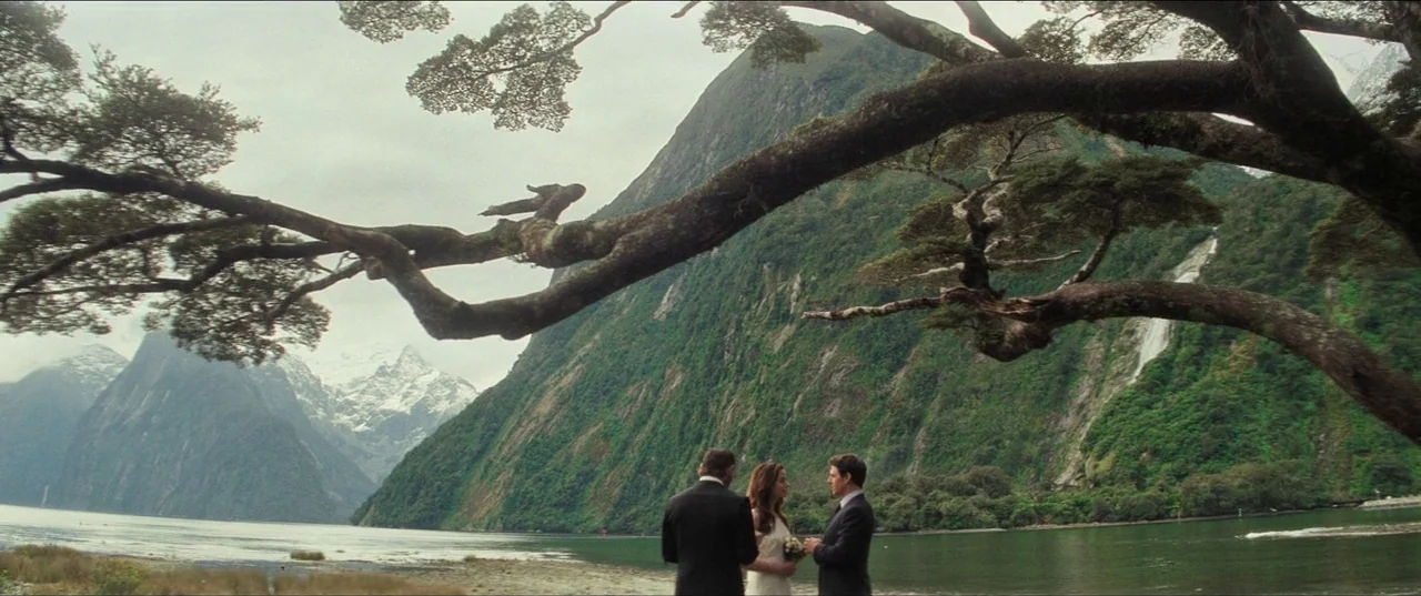 A man and woman stand facing each other before a man in a suit. They are on the shore of a vast lake framed by dramatic steep mountain slopes.