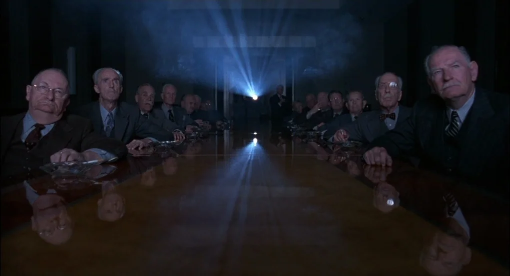 The Hudsucker Proxy (1994) movie still: wide — A dark boardroom is occupied by rows of elderly men seated at a long table. They watch a beam of…