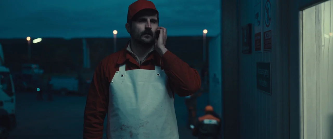 R.M.N. (2022) movie still: medium — An industrial outdoor loading dock at night features signage in German on a white wall. A man in a red uniform…