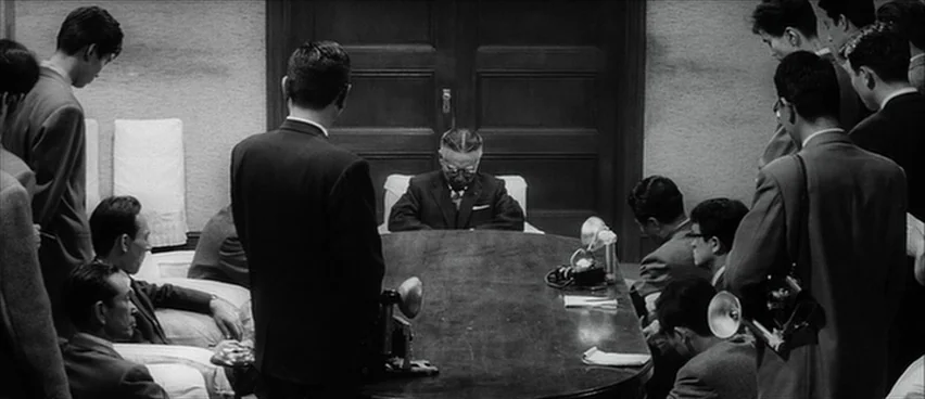 The Bad Sleep Well (1960) movie still: wide — A formal room contains a long, dark, polished oval wooden table. Multiple men in suits gather around…
