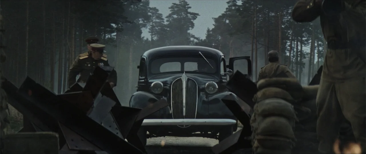 Sisu: Road to Revenge (2025) movie still: wide — A dark vintage car sits parked on a dirt road surrounded by pine trees. Two military officers in…