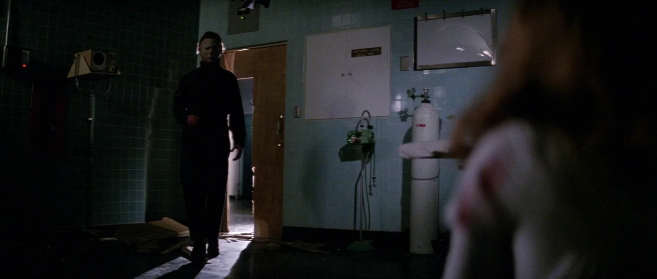 Halloween II (1981) movie still: medium — A dark hospital room features teal tiled walls and medical equipment in the background. A man wearing a…