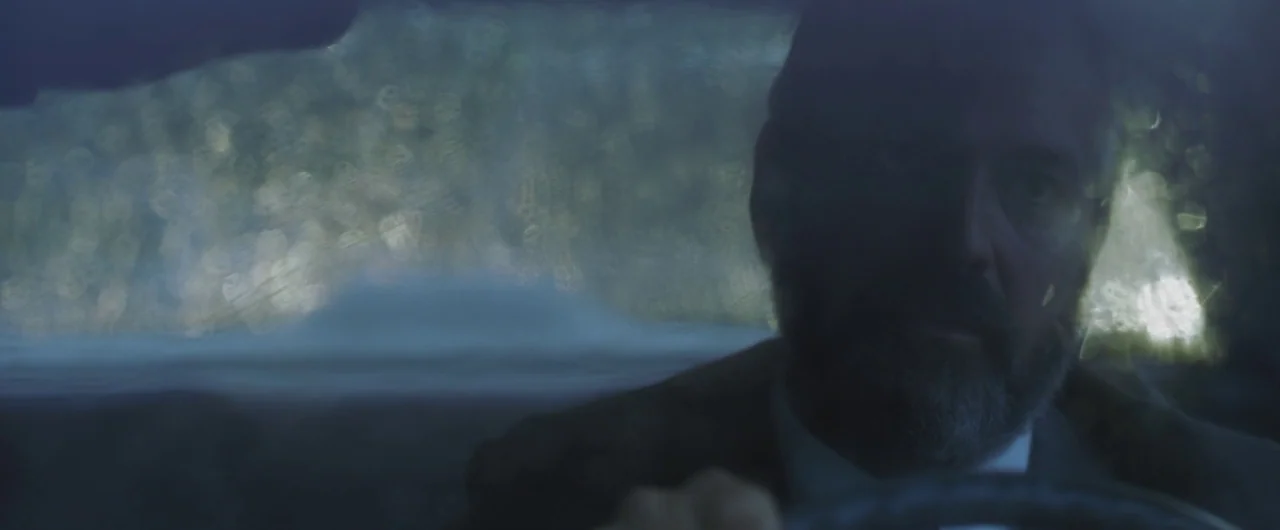 Syriana (2005) movie still: closeup — An interior of a car at night with rain streaking the glass window pane. A man with a dark beard sits in the…