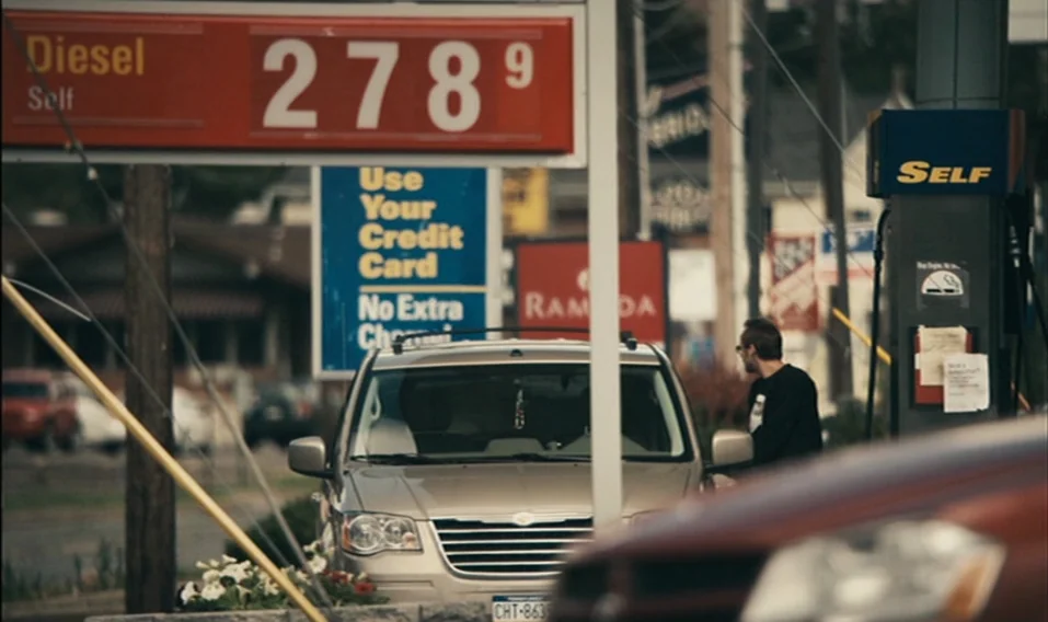Blue Valentine (2010) movie still: medium — A gas station exterior features a large red pricing sign with diesel costs displayed. A man stands…