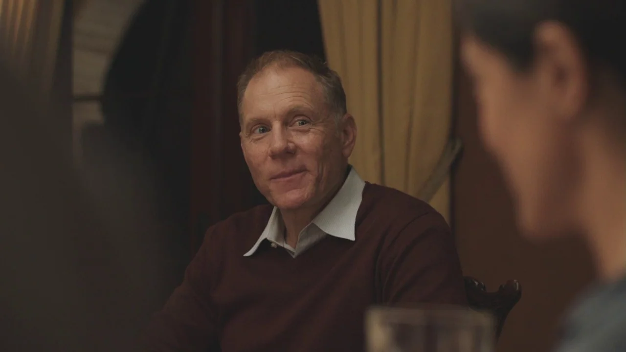 Beatriz at Dinner (2017) movie still: medium — A dimly lit room with draped curtains visible in the background. A middle-aged man in a burgundy…