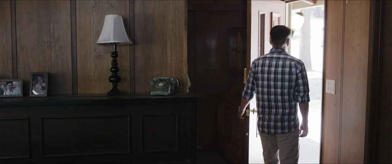 Captive (2020) movie still: medium — A wood-paneled residential hallway features a vintage green rotary phone and a table lamp. A man stands at an…