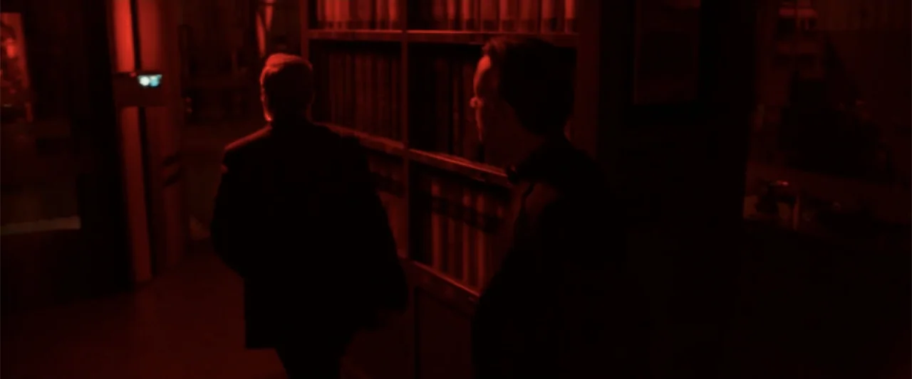 Angels & Demons (2009) movie still: medium — A dark, dimly lit library corridor features floor-to-ceiling shelves packed with books. Two men in dark…