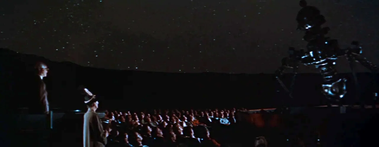 Rebel Without A Cause (1955) movie still: wide — A dark planetarium auditorium has a large mechanical star projector positioned to the right. A…