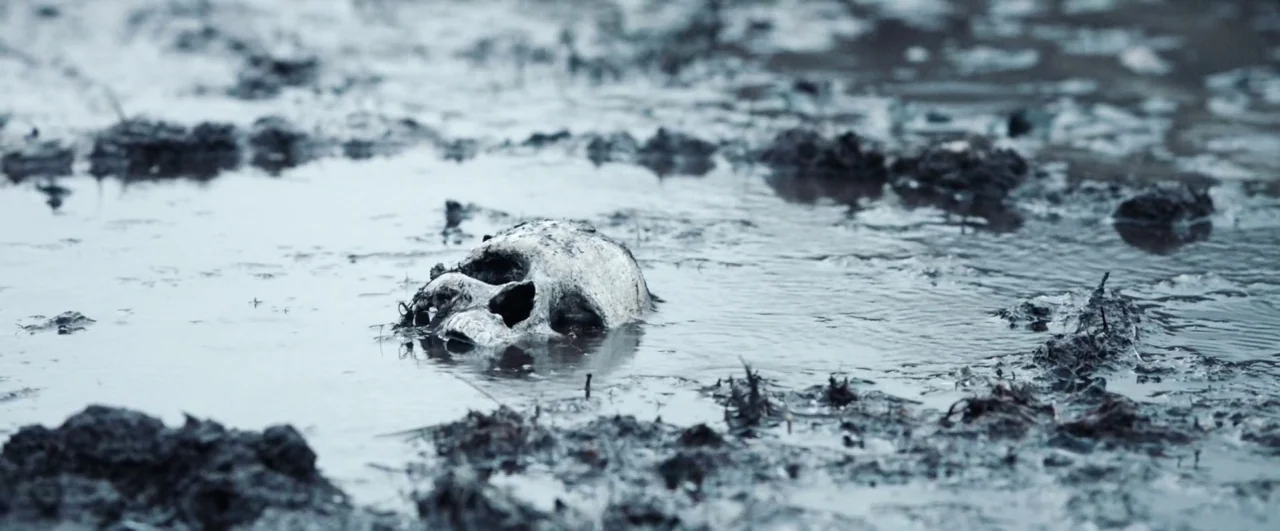Black ’47 (2018) movie still: closeup — A human skull sits half-submerged in a shallow pool of murky water. The surrounding ground consists of dark,…