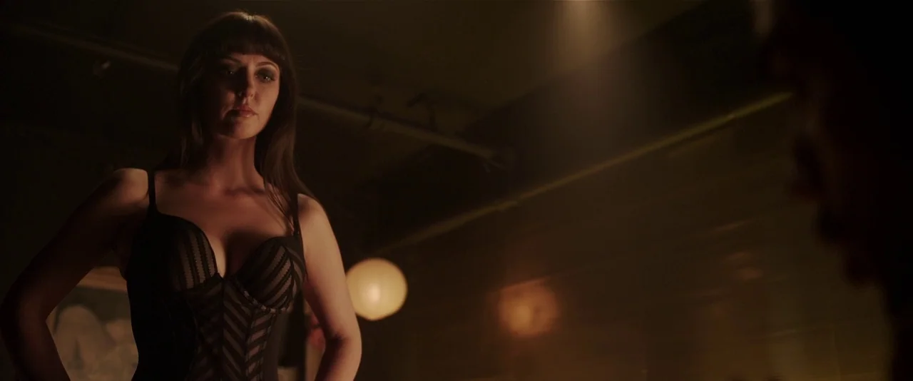 American Mary (2012) movie still: medium — A dimly lit room with an overhead light fixture illuminating a person. A woman stands in the foreground…