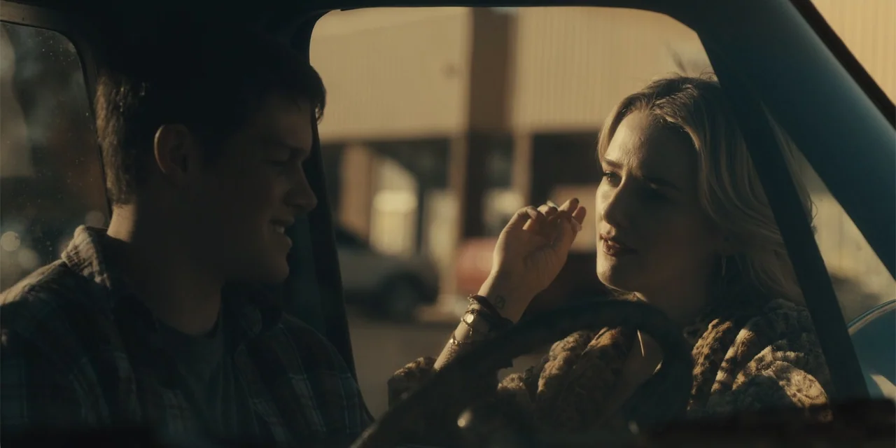 All Roads to Pearla (2019) movie still: medium — Two individuals sit in the front seats of a vehicle during the late afternoon. A man and a woman…