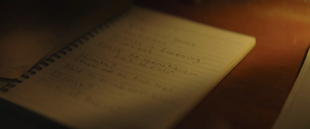 Wolf Man (2025) movie still: closeup — A notebook with spiral binding lies open on a dimly lit wooden desk surface. Handwritten notes in pencil fill…