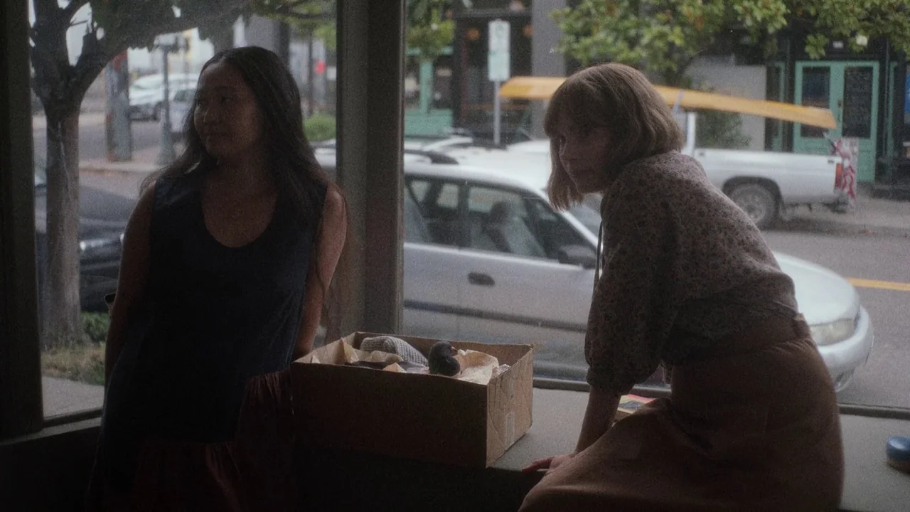Showing Up (2022) movie still: medium — Two women stand inside a storefront looking out through a large window onto the street. A cardboard box…