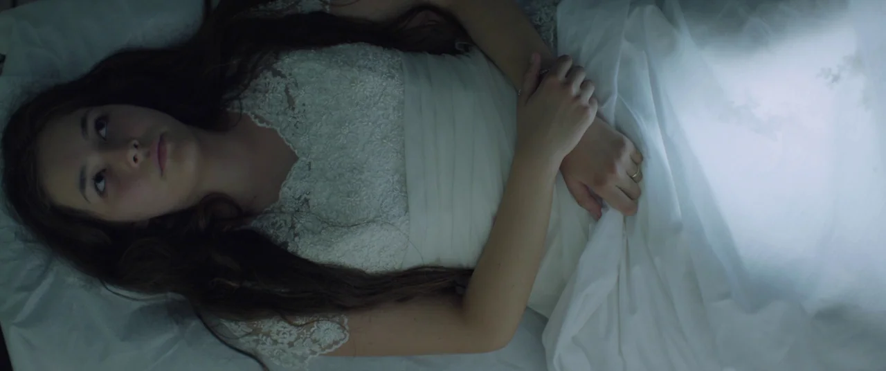 Mustang (2015) movie still: medium — A woman lies on white bed sheets wearing a formal white lace gown. She remains still with her arms folded…