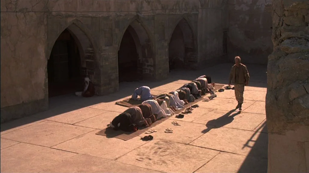 The Exorcist (1973) movie still: wide — A stone-walled courtyard with arched doorways features prayer rugs laid out on the ground. A group of men…