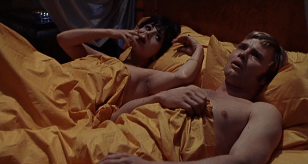 Midnight Cowboy (1969) movie still: medium — A dark, cramped room featuring a bed with bright yellow linens. A man and a woman lie together while…