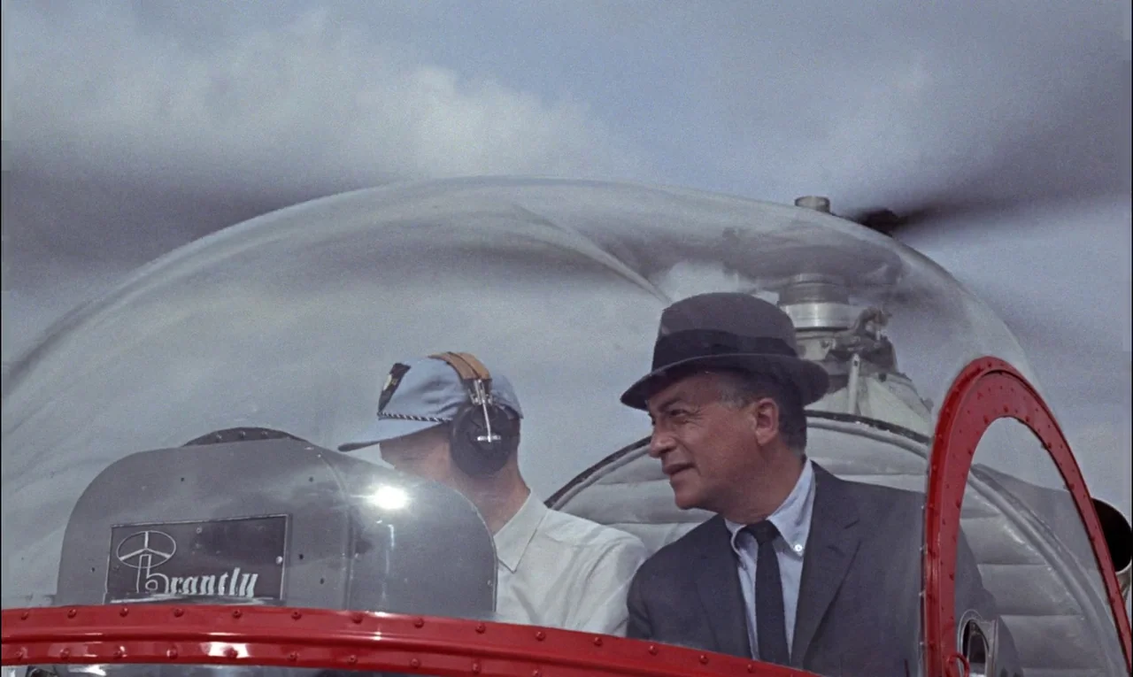 Goldfinger (1964) movie still: medium — Two men sit inside the clear bubble canopy of a small Brantly helicopter. The pilot operates controls while…
