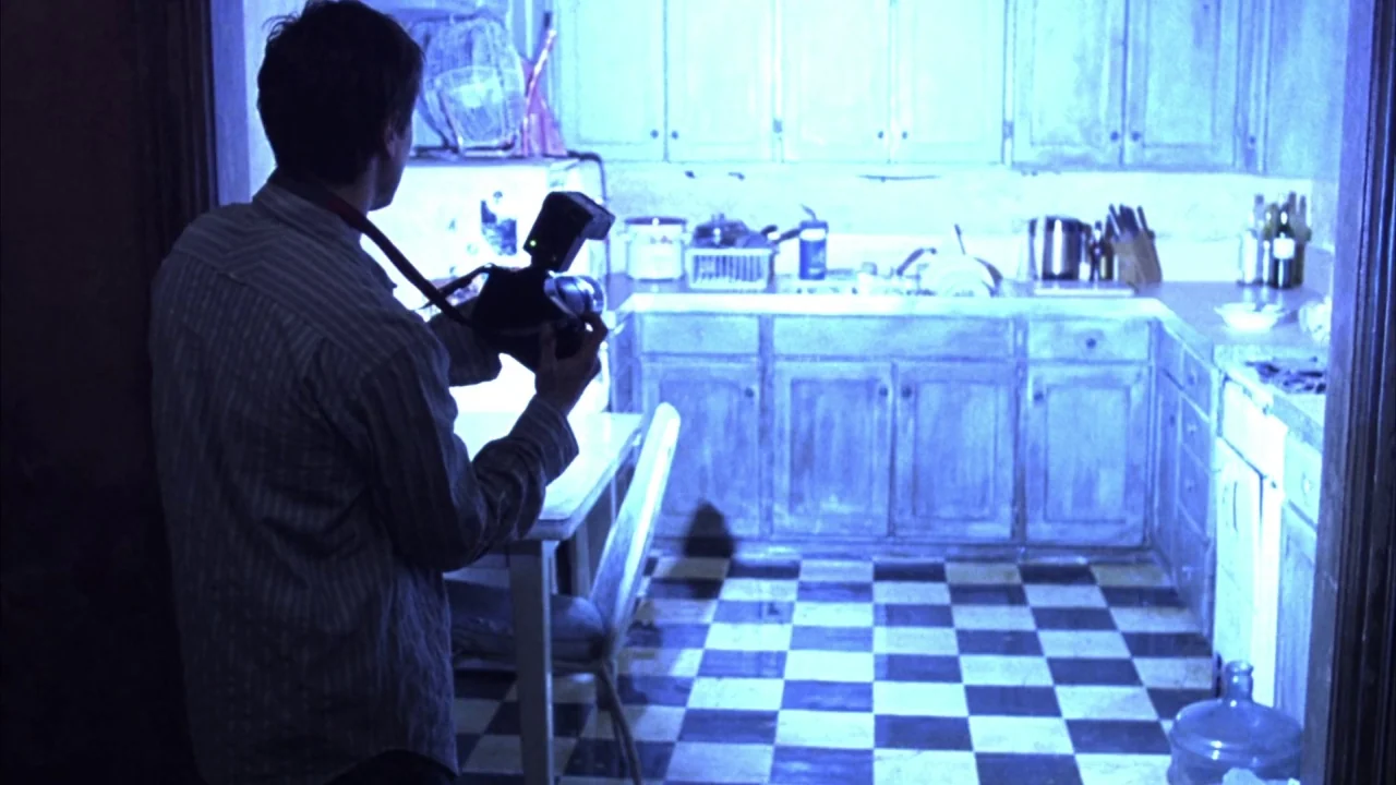 Saw movie still: medium — A residential cooking space features black and white checkerboard floor tiles and wood cabinetry. A man stands in the…