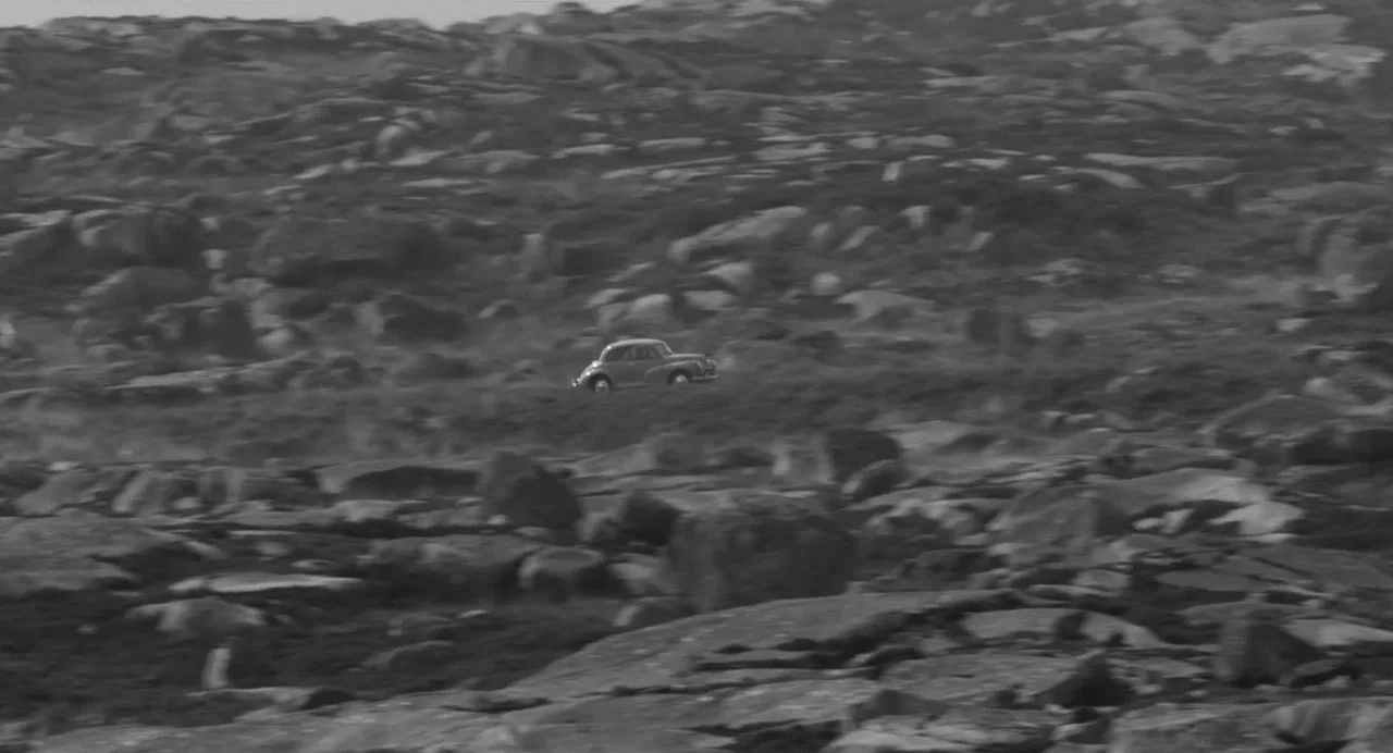 Song of Granite (2017) movie still: wide — A vast field covered in large jagged rocks stretches toward a distant horizon line. A small vintage car…